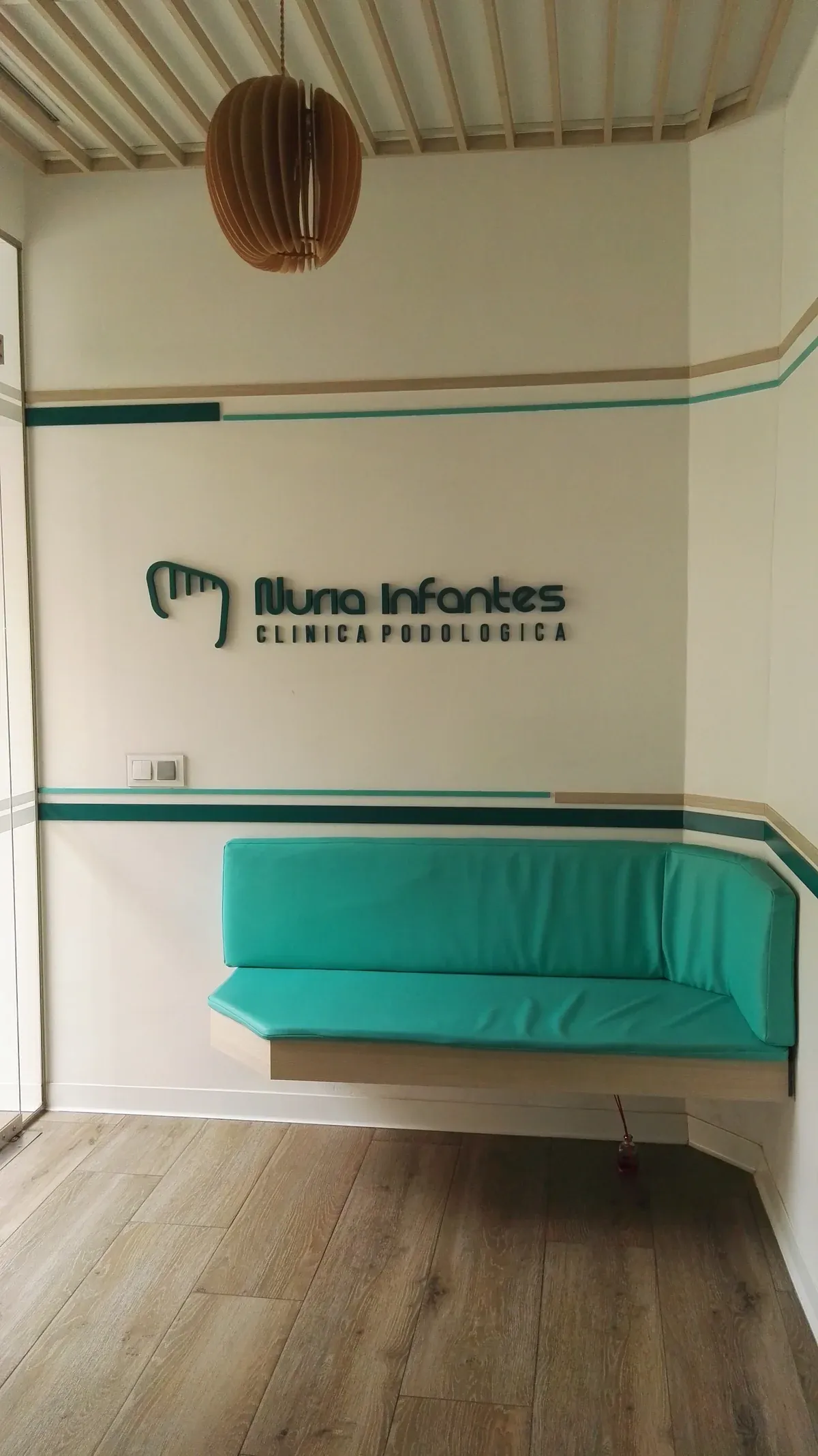 Sala de espera de la Clínica Podológica Nuria Infantes en Daimiel: sofá turquesa, rótulo corporativo y techo de listones.